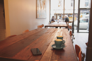 The image features a wooden table with coffee cups on it, with a large storefront window in the distance.